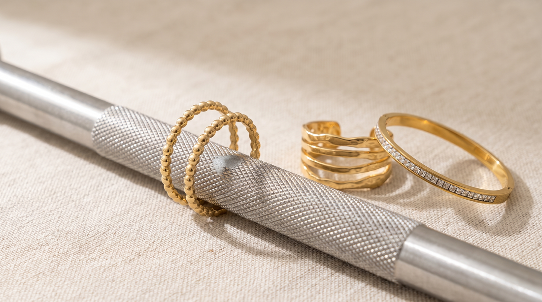 Goldfarbener offener Edelstahlring liegt auf der Rändelung einer Hantelstange, an der Berührungsstelle ist die Vergoldung bis auf graues Metall durchgerieben; daneben ein unbeschädigter Ring und ein Armreif.