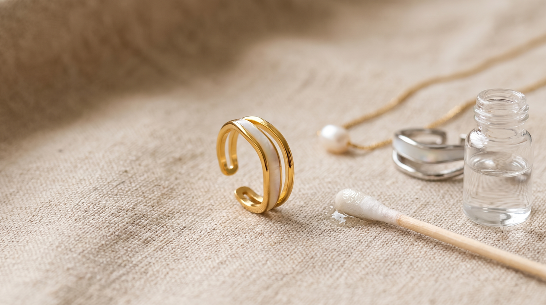 Goldfarbener Ring mit Perlmutt-Einlage auf beigem Leinen, daneben ein feuchtes Wattestäbchen und ein Fläschchen mit klarer Flüssigkeit; rechts ein blanker Edelstahlring und eine Kette mit Perle.