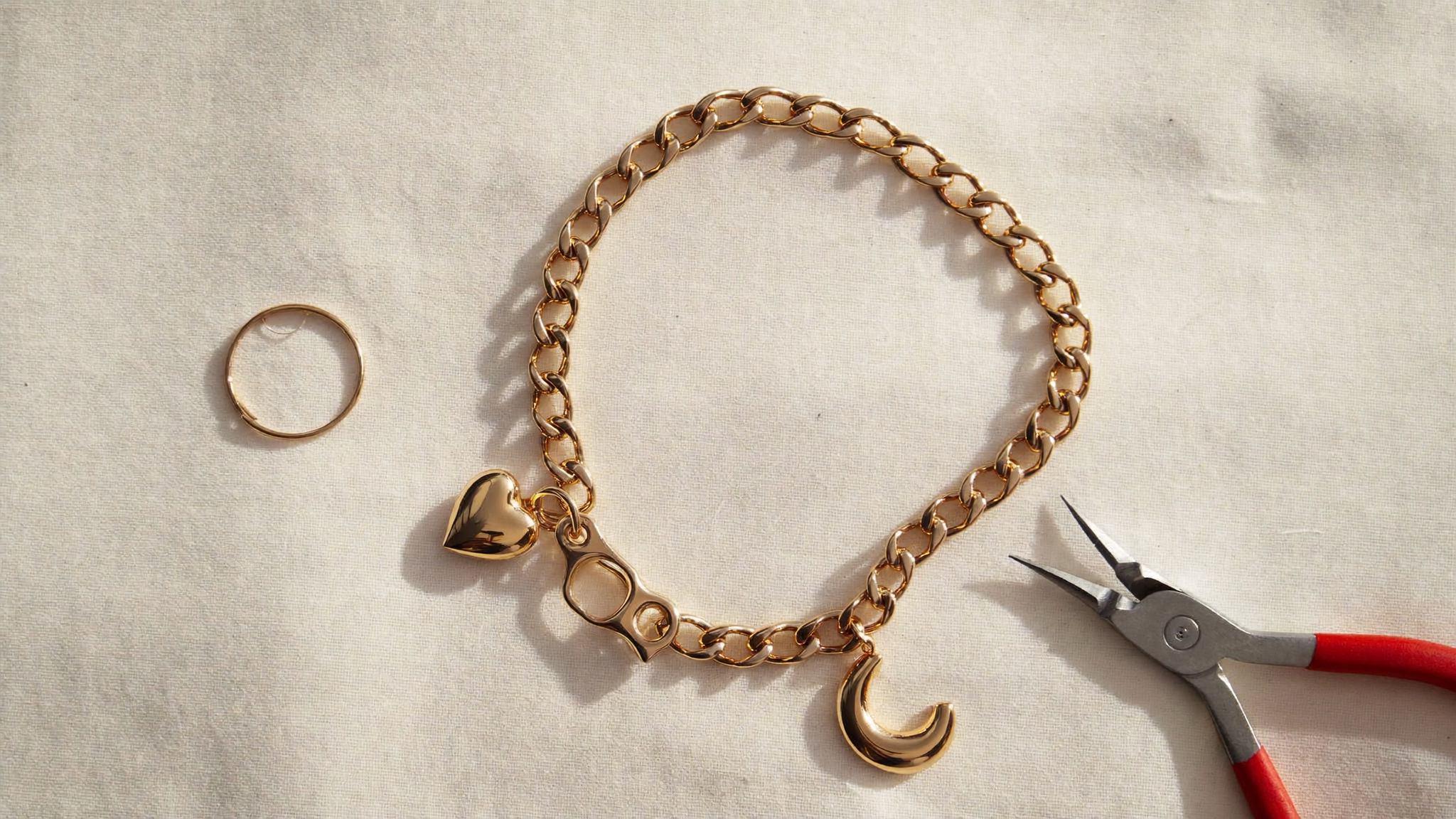 Goldfarbenes Gliederarmband aus Edelstahl mit Herz-Charm und Mond-Charm flach auf Leinen ausgelegt, daneben eine Flachzange und ein offener Biegering
