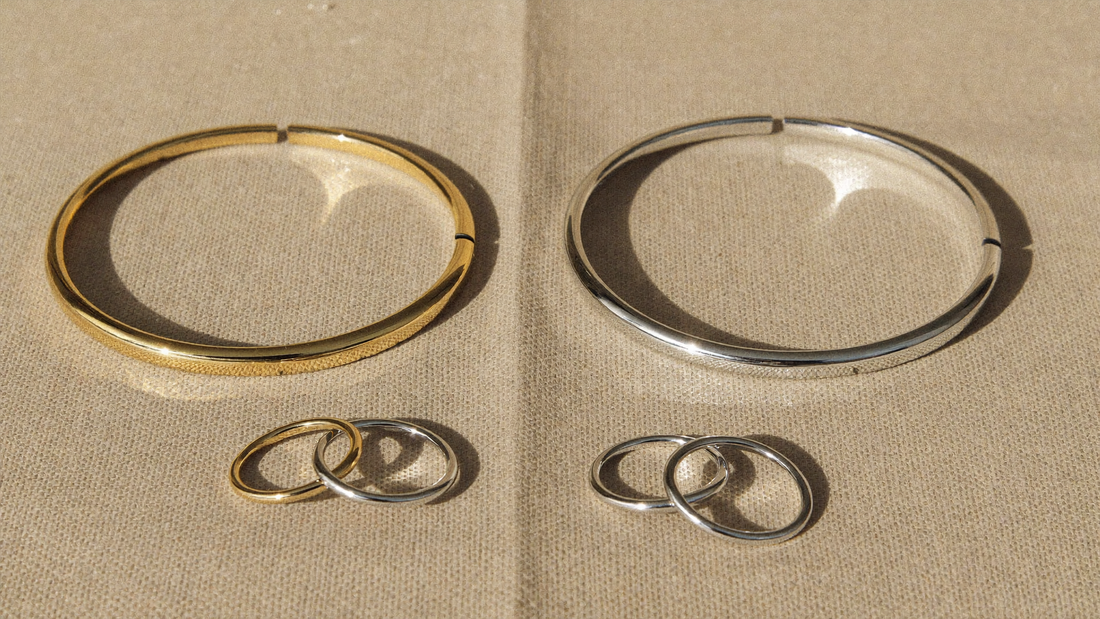 Ein goldfarbener und ein silberfarbener Armreif aus Edelstahl liegen nebeneinander auf beigem Leinen, davor Ringe in Gold und Silber