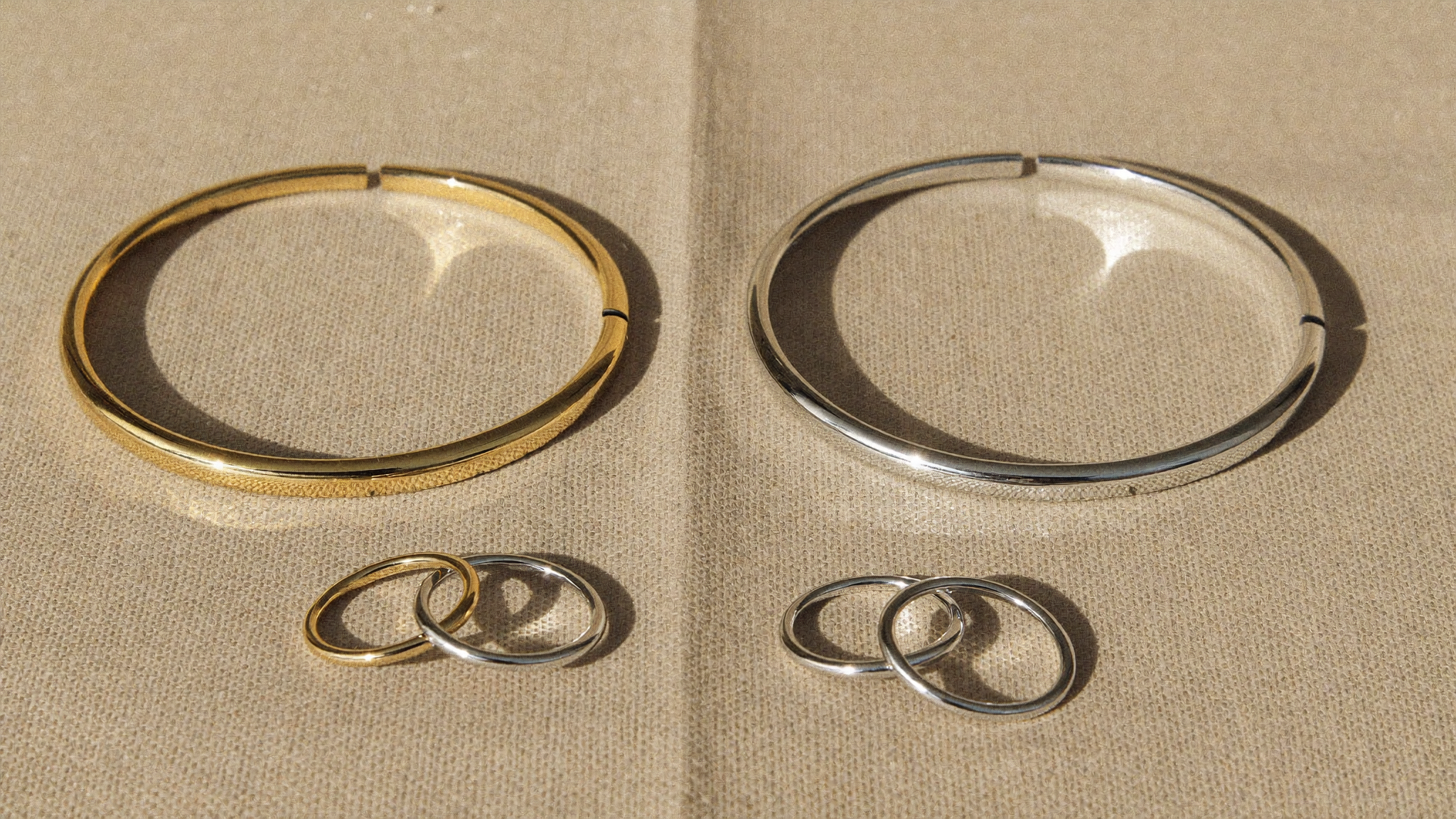 Ein goldfarbener und ein silberfarbener Armreif aus Edelstahl liegen nebeneinander auf beigem Leinen, davor Ringe in Gold und Silber